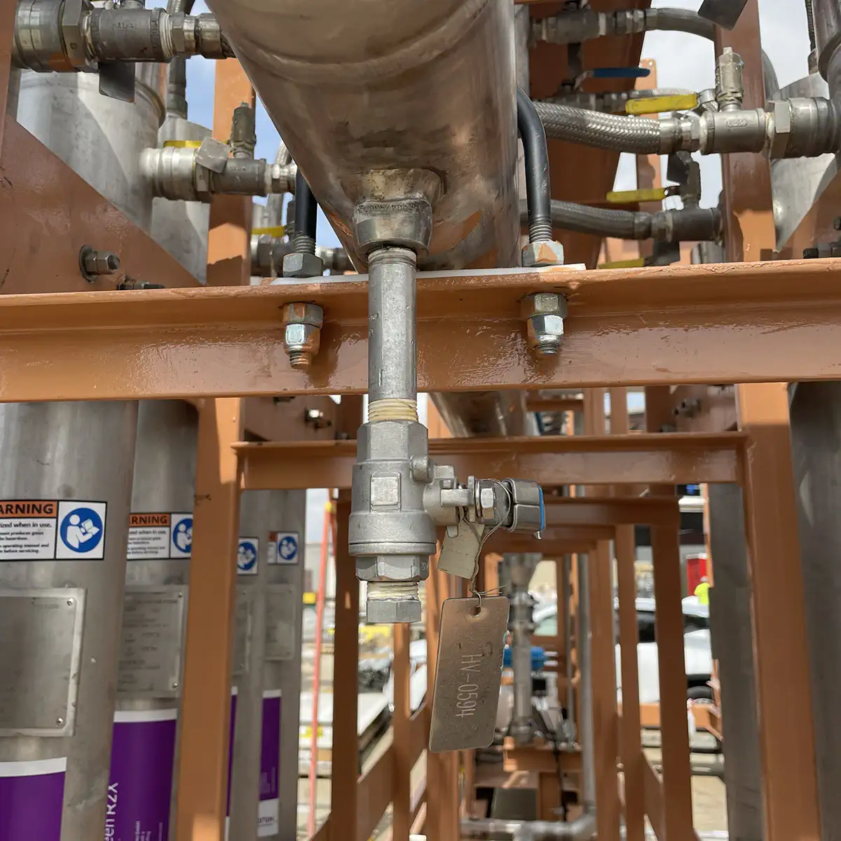 Industrial piping with a tagged valve on an orange beam.