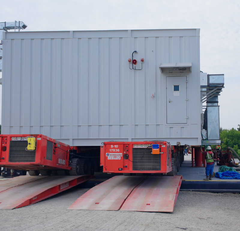 Prefabricated industrial module positioned on heavy transport equipment beside a barge during loading operations.